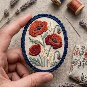 May include: An oval-shaped embroidered patch featuring a floral design of red and dark orange poppies with green stems and cream-colored leaves on a light beige fabric background, finished with a dark blue scalloped edge trim border.