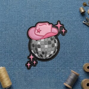 May include: A patch featuring a pink cowboy hat with a star detail resting on a silver disco ball, accented by small pink four-pointed stars. The design is outlined in black and set against a textured blue fabric background with sewing supplies.