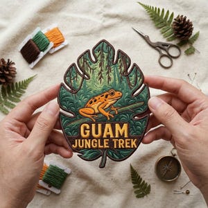 May include: An embroidered patch shaped like a monstera leaf featuring a bright orange frog perched on a branch. The design includes dark green foliage and the text GUAM JUNGLE TREK in bold yellow letters. The patch has a dark brown stitched border.