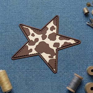 May include: A star-shaped patch featuring a brown and cream cow print pattern. The patch has a dark brown border with visible stitching along the inner edge, creating a defined outline for the star shape against the textured blue fabric background.