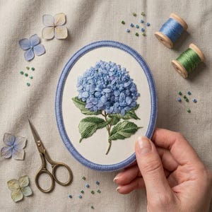 May include: An oval embroidery hoop containing a fabric panel with a detailed blue hydrangea flower and green leaves. The hoop has a blue border. Nearby are two wooden spools of blue and green thread, small scissors, and loose beads.