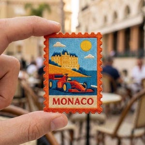 May include: A rectangular embroidered patch with a scalloped orange border featuring a red race car on a blue track, a yellow building on a hill, a yellow sun, and white clouds against a blue sky, with the word MONACO printed in red letters.