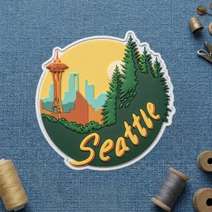 Puede incluir: Una pegatina redonda con el horizonte de Seattle, incluyendo la Space Needle, árboles y la palabra "Seattle" en amarillo. La pegatina tiene un borde verde y un fondo amarillo y naranja.