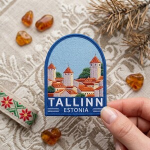 May include: A rectangular embroidered patch with a rounded top edge featuring a detailed scene of the Tallinn, Estonia skyline with orange-roofed towers against a light blue sky. The text TALLINN ESTONIA is stitched in white at the bottom.