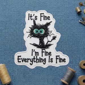 May include: A white patch with a black cat illustration and the text "It's Fine, I'm Fine, Everything Is Fine." The cat has large, teal eyes and a distressed appearance. The patch is on a blue textured surface.