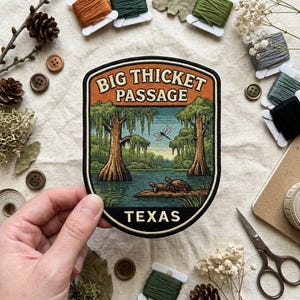 May include: An embroidered patch featuring a landscape of the Big Thicket Passage in Texas. The design shows two turtles on a log in a river, cypress trees, and a dragonfly, with the text BIG THICKET PASSAGE and TEXAS in cream lettering.