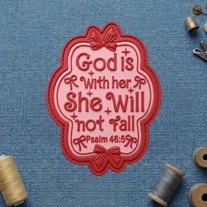 May include: A pink and red decorative plaque with the text "God is with her, She Will not Fall" and "Psalm 46:5". The plaque has a bow at the top and bottom. The background is blue fabric.