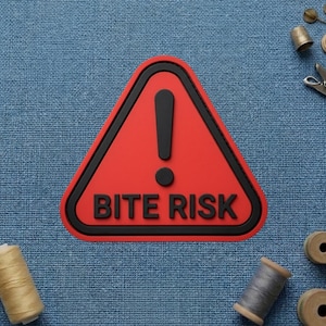 May include: A red triangular warning sign with a black border and the words "BITE RISK" in black. The sign features a black exclamation point. Sewing supplies, including spools of thread and a thimble, are scattered around the sign on a blue fabric background.