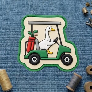 May include: A whimsical patch featuring a white duck driving a green golf cart. The cart has black wheels and a red golf bag. The patch has a green border and a beige background.