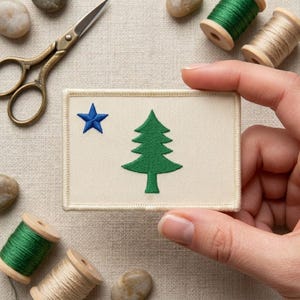 May include: A rectangular fabric patch with a cream-colored background featuring an embroidered green pine tree in the center and a small blue star in the upper left corner. The patch has a stitched border and is held against a neutral fabric surface.