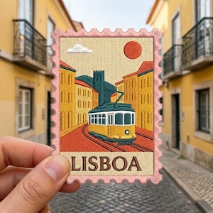 May include: A rectangular embroidered patch shaped like a postage stamp with a pink scalloped border. It features a yellow tram on tracks between yellow buildings, a red sun, and a white cloud. The text LISBOA is embroidered in brown at the bottom.