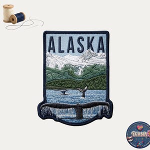 May include: A rectangular embroidered patch featuring the text ALASKA above a landscape of snow-capped mountains, green forests, and blue water with whale tails breaching the surface. Two wooden spools of thread and a needle are included.