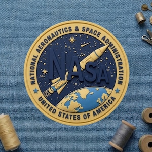 May include: A round NASA emblem with a blue and yellow color scheme. The emblem features a rocket, stars, and the words "National Aeronautics & Space Administration" and "United States of America". The NASA logo is in the center.