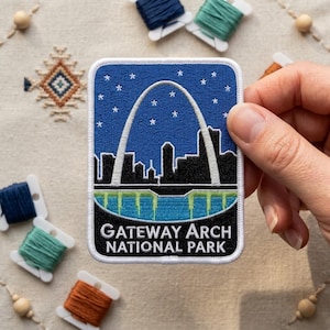 May include: A rectangular embroidered patch featuring a blue night sky with white stars, a white Gateway Arch silhouette, a black city skyline, and a green river. The text GATEWAY ARCH NATIONAL PARK is embroidered in white at the bottom.