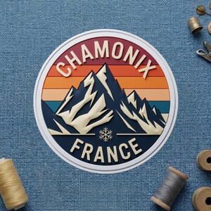May include: A round sticker featuring a mountain range design with the text "CHAMONIX FRANCE". The design includes a color gradient of orange, red, and blue. A snowflake is also included in the design.