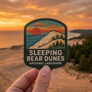 May include: A shield-shaped embroidered patch featuring a landscape of Sleeping Bear Dunes National Lakeshore. The design shows a red sun over a blue lake, a large sand dune, and green pine trees. The text reads SLEEPING BEAR DUNES NATIONAL LAKESHORE.
