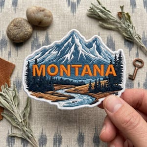 May include: An embroidered patch featuring a mountain landscape with snow-capped peaks, a winding river, and evergreen trees. The word MONTANA is stitched in bold orange letters across the center of the design, which is outlined in white thread.