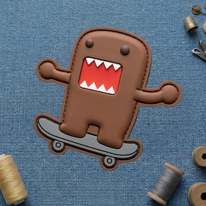 May include: A brown Domo-kun patch on a skateboard, with a red mouth and white teeth. The patch is brown with a gray skateboard. The background is a blue fabric.