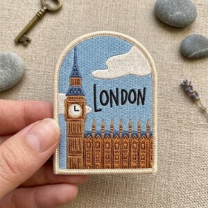 May include: An arched embroidered patch featuring a light blue sky with white clouds and the brown Elizabeth Tower, commonly known as Big Ben, alongside the Houses of Parliament. The word LONDON is stitched in black thread above the building.