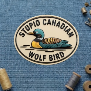 May include: An oval-shaped patch with a cartoon illustration of a loon. The loon has a yellow chest, teal wings, and a black and white neck. The text "STUPID CANADIAN WOLF BIRD" is in black, around the loon.