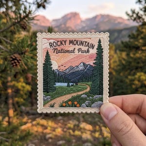 May include: A rectangular embroidered patch with a scalloped edge featuring a landscape of Rocky Mountain National Park. The design includes tall pine trees, a winding dirt path, a lake, mountains, and the text Rocky Mountain National Park.