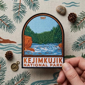 May include: An embroidered patch featuring a landscape of Kejimkujik National Park with a river, orange rock formations, and green pine trees under a light blue sky. The text 1967 is at the top, and KEJIMKUJIK NATIONAL PARK is at the bottom.