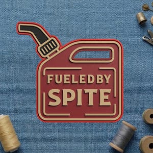 May include: A red and tan gas can-shaped design with the words "FUELED BY SPITE" in a bold font. The design has a black nozzle and is set against a blue fabric background. Sewing notions are scattered around the design.