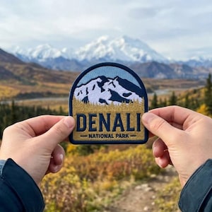 May include: A semi-circular embroidered patch featuring a mountain landscape with a light blue sky, white snow-capped peaks, and a golden-brown forest. The text DENALI NATIONAL PARK is stitched in dark blue lettering across the lower section.