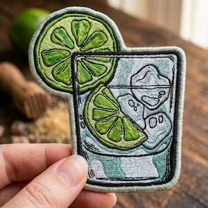 May include: An embroidered patch featuring a cocktail glass filled with ice cubes and a lime wedge, with a large lime slice positioned behind the top of the glass. The design uses shades of green, white, and light blue thread on a fabric base.