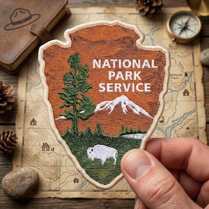 May include: An embroidered National Park Service patch in the shape of an arrowhead. The patch features a brown landscape with a green tree, a white snow-capped mountain, and a white bison. The text NATIONAL PARK SERVICE is stitched in white.