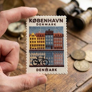 May include: A rectangular embroidered patch shaped like a postage stamp featuring a cityscape of KØBENHAVN DENMARK. The design shows colorful buildings in yellow, blue, and brown, a black bicycle, and the text KØBENHAVN DENMARK on a cream background.