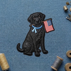 May include: A black Labrador retriever patch featuring a detailed dog sitting upright, wearing a light blue collar with a bow, and holding a small American flag in its mouth. The design is rendered in a raised, textured style with clear outlines.