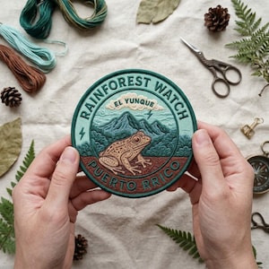 May include: A circular embroidered patch featuring a brown frog in the foreground, green mountains, and a cloud with the text "EL YUNQUE" above. The border text reads "RAINFOREST WATCH" and "PUERTO RICO" in dark green on a light green background.