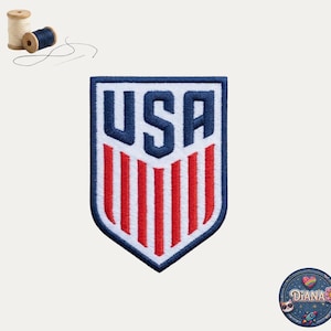 May include: A shield-shaped embroidered patch featuring the text USA in dark blue block letters above five vertical red stripes on a white background, bordered in dark blue, accompanied by two small spools of thread and a needle.