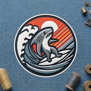 May include: A round, colorful patch featuring a cartoon shark surfing a wave. The design includes a red and orange sunset, white sun, and a blue and white wave. The shark is gray and white, with a black outline.