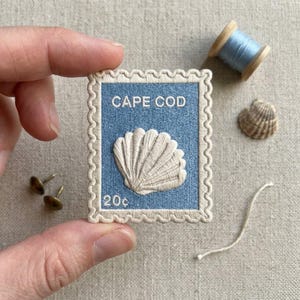 May include: A rectangular embroidered patch shaped like a postage stamp with a light blue background and a cream-colored scalloped border. It features an off-white seashell graphic, the text CAPE COD, and the denomination 20c in white thread.