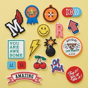 May include: A collection of colorful, decorative patches with various designs and text. Includes a lightning bolt, a smiley face, a compass, a pizza inspector badge, and patches with the words "AMAZING", "YOU ARE AWESOME", and "Try it! You'll like it!"