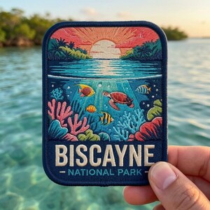 May include: A rectangular embroidered patch featuring a sunset over water with a coral reef scene below. The design includes a sea turtle, tropical fish, and coral in shades of pink, blue, and orange. The text reads BISCAYNE NATIONAL PARK.