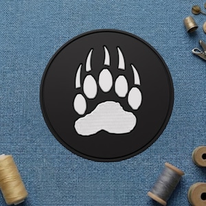 May include: A round, black patch featuring a white bear paw print. The paw print has five toes and a textured sole. The patch is likely made of a durable material and is suitable for sewing or ironing onto clothing or accessories.