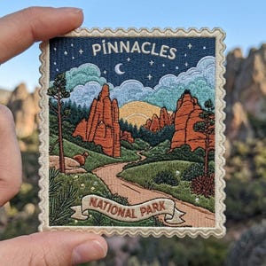 May include: A rectangular embroidered patch with a scalloped edge featuring a landscape of Pinnacles National Park. The design shows orange rock formations, a winding path, green trees, and a night sky with a crescent moon and stars.