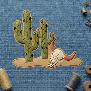 May include: A desert-themed patch featuring two green cacti with black spines and a cow skull with tan horns. The patch is set on a tan base, with a blue background.