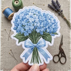 May include: An embroidered patch featuring a bouquet of light blue hydrangea flowers with green leaves and stems, tied together with a light blue ribbon. The patch has a white scalloped border and is shaped to follow the outline of the floral design.