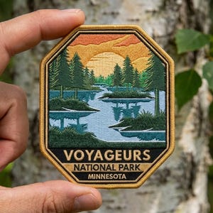 May include: An octagonal embroidered patch featuring a landscape of Voyageurs National Park in Minnesota. The design shows a blue lake, green pine trees, and a yellow sunset. The text reads VOYAGEURS NATIONAL PARK MINNESOTA in gold letters.