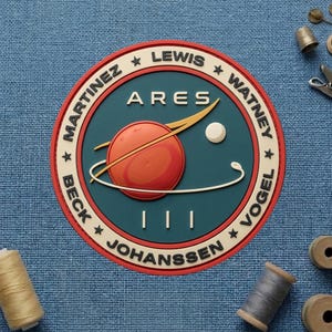 May include: A circular patch with a red planet and the word "ARES" in white. The patch has a teal background with the names of crew members around the edge. The patch is on a blue fabric surface with sewing supplies.