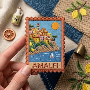 May include: A rectangular embroidered patch with a scalloped border featuring a coastal scene of Amalfi. The design includes colorful buildings on a cliff, a yellow sun, a sailboat on blue water, and the text AMALFI in a tan banner at the bottom.