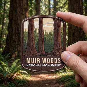 May include: A rectangular embroidered patch with rounded corners featuring a forest scene of tall brown redwood trees against a light tan sky. The text MUIR WOODS NATIONAL MONUMENT is stitched in white letters on a dark brown background.