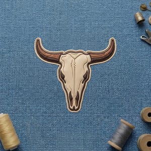 May include: A bull skull patch featuring a cream-colored cranium with dark brown accents and curved, textured brown horns. The design is rendered in a flat, graphic style with clean outlines, showcasing a symmetrical shape and detailed horn shading.