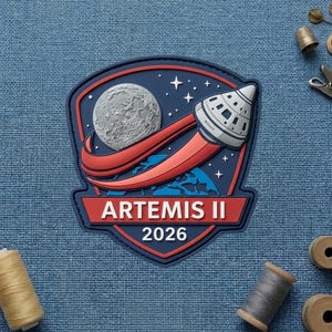 May include: A blue, red, and white Artemis II 2026 patch. The patch features a detailed illustration of the moon, a rocket, and the Artemis II logo. The patch is on a blue fabric background.
