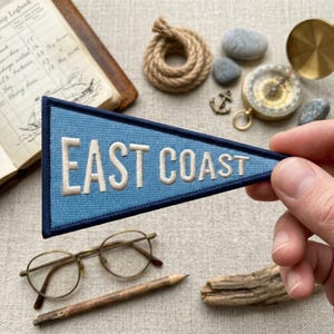May include: A triangular blue felt pennant with a dark blue border featuring the embroidered white text EAST COAST. The pennant is held against a background containing a vintage logbook, a coil of rope, a brass compass, and eyeglasses.