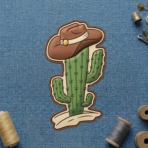 May include: A cartoon-style cactus wearing a brown cowboy hat with a white band and gold buckle. The cactus is green with small white star-shaped details. The design has a brown border and sits on a beige base.